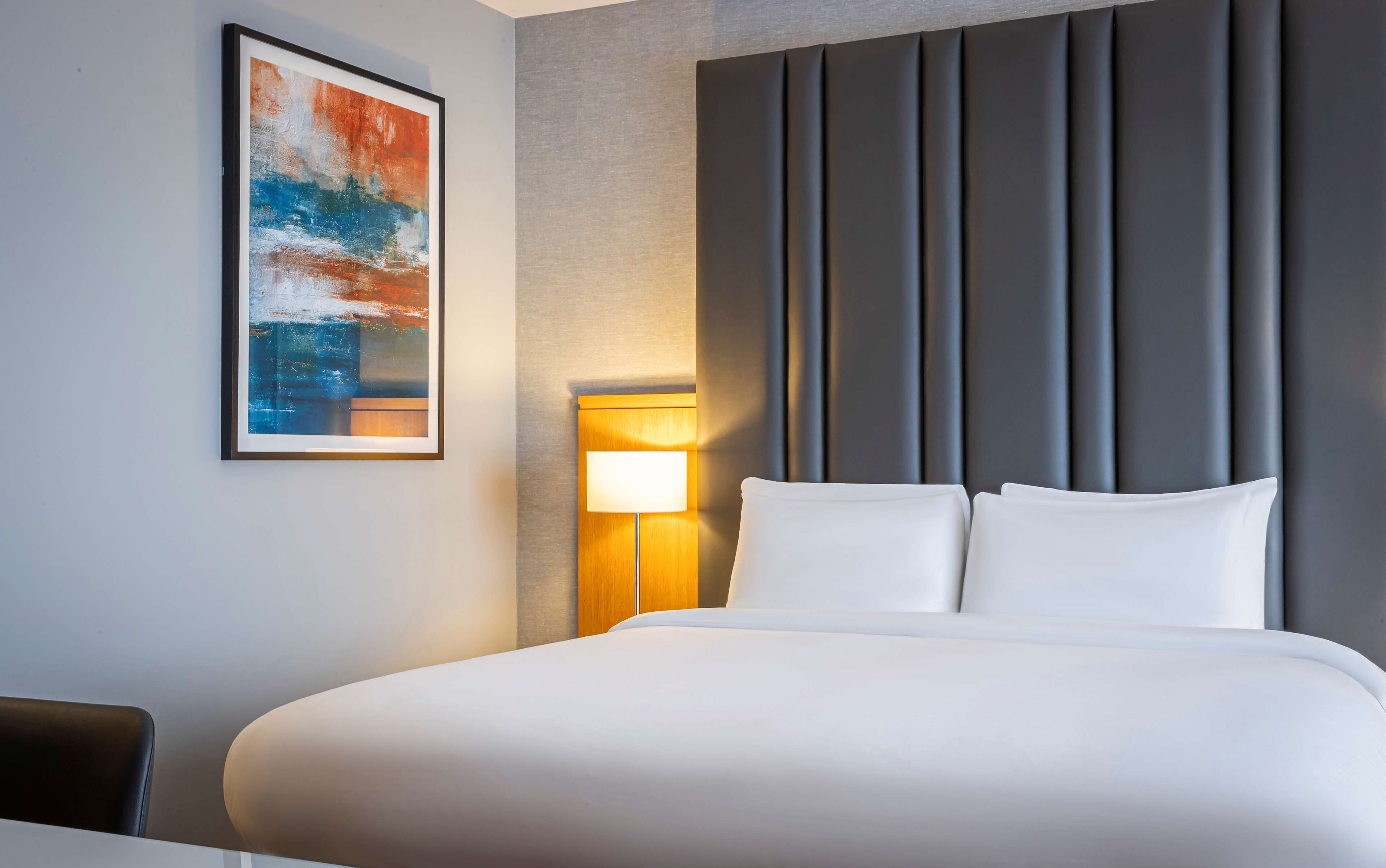 A hotel room with a large bed, a headboard with vertical slats, a wall-mounted lamp, and a framed abstract painting.