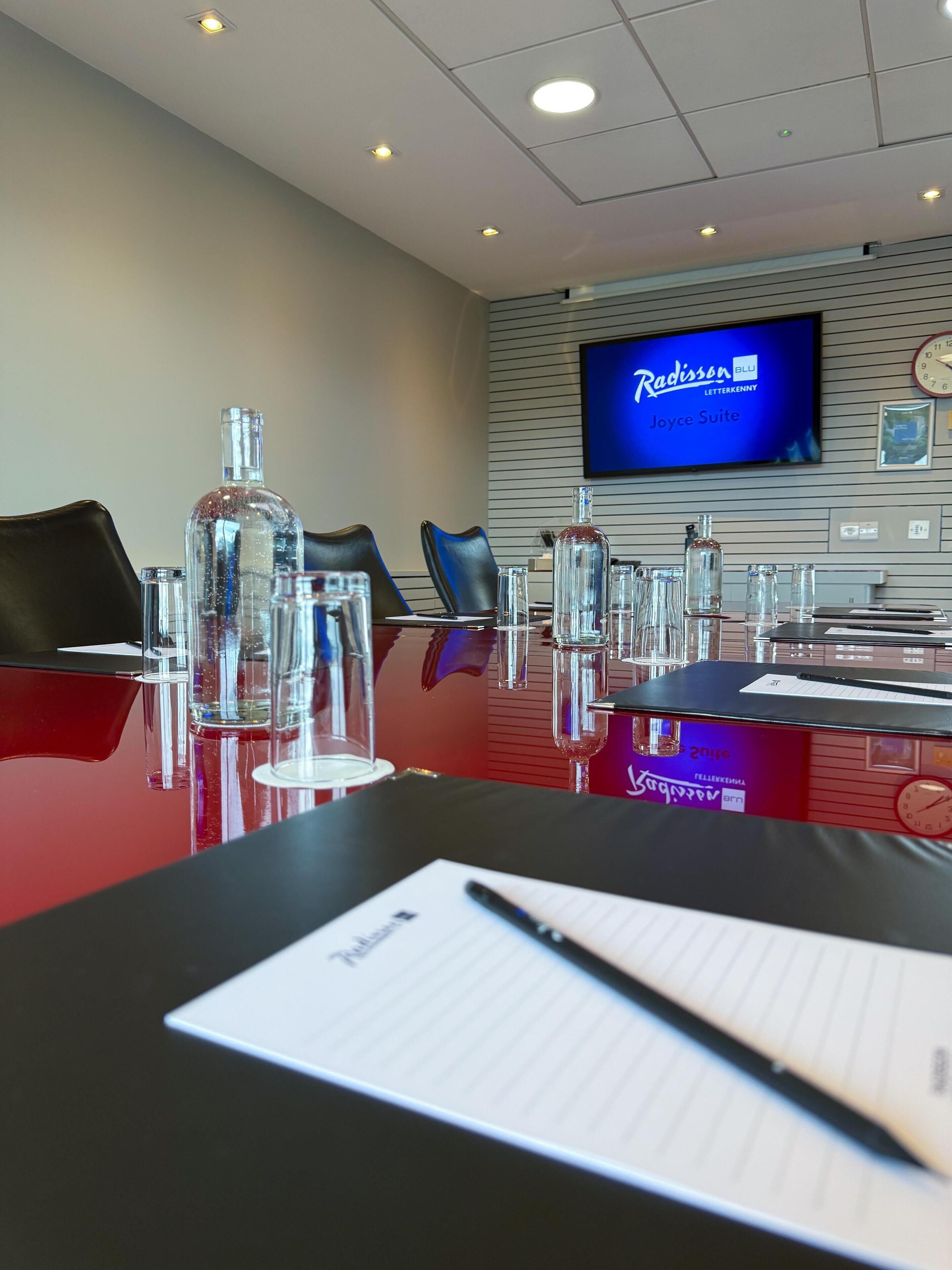 A conference room with a glass table, chairs, and a television displaying 'Radisson Blu Joyce Suite'.