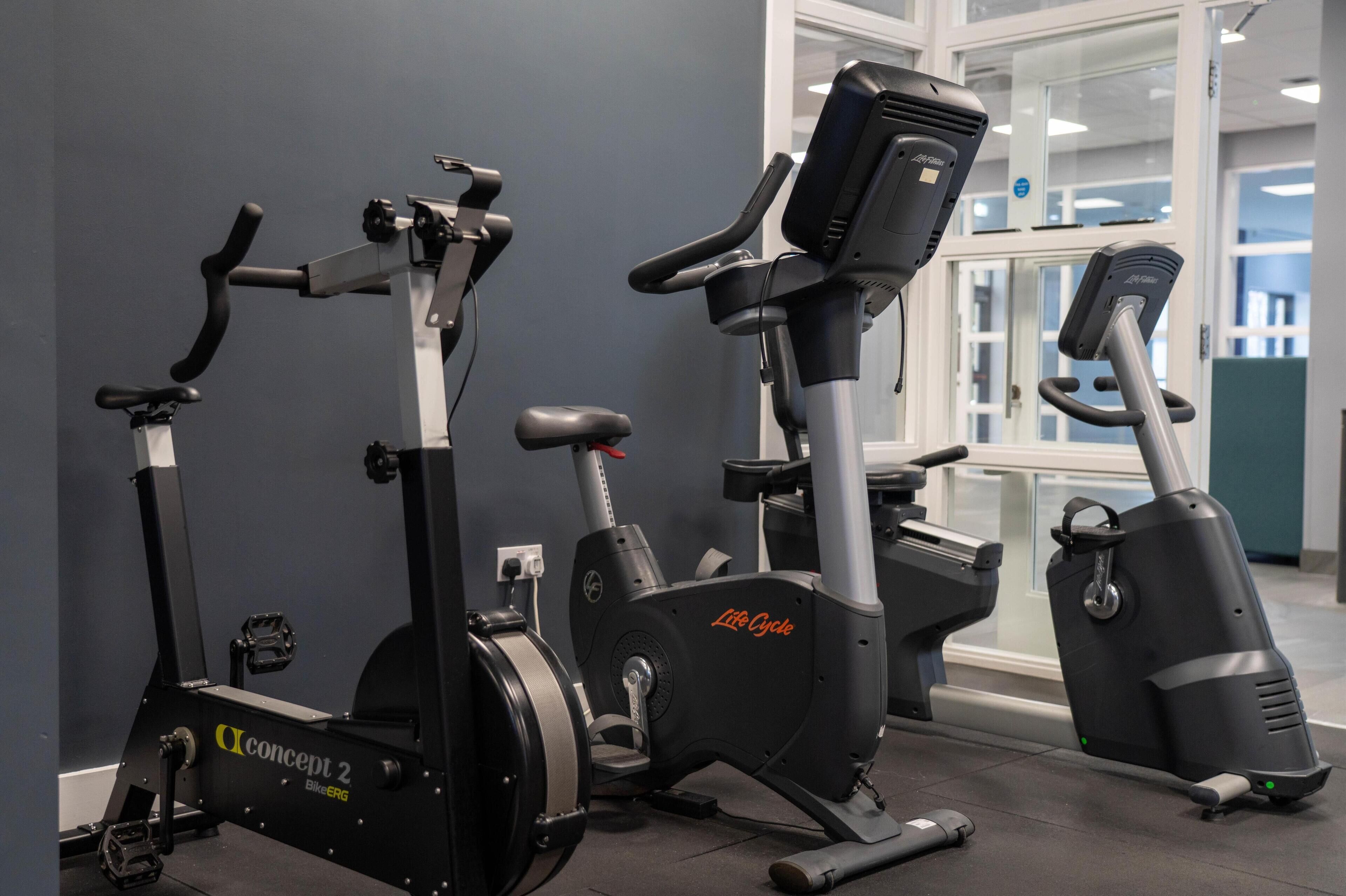 Tres bicicletas estáticas en un gimnasio con una pared gris y una ventana al fondo.