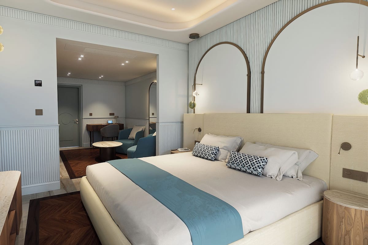 Una habitación de hotel con una cama grande, una zona de estar con una mesa y sillas, un espejo y vista a la habitación contigua.