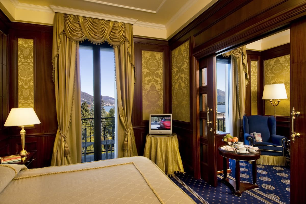 Quarto de hotel com cama, televisão, mesinha com abajur, cadeira e vista para o exterior.