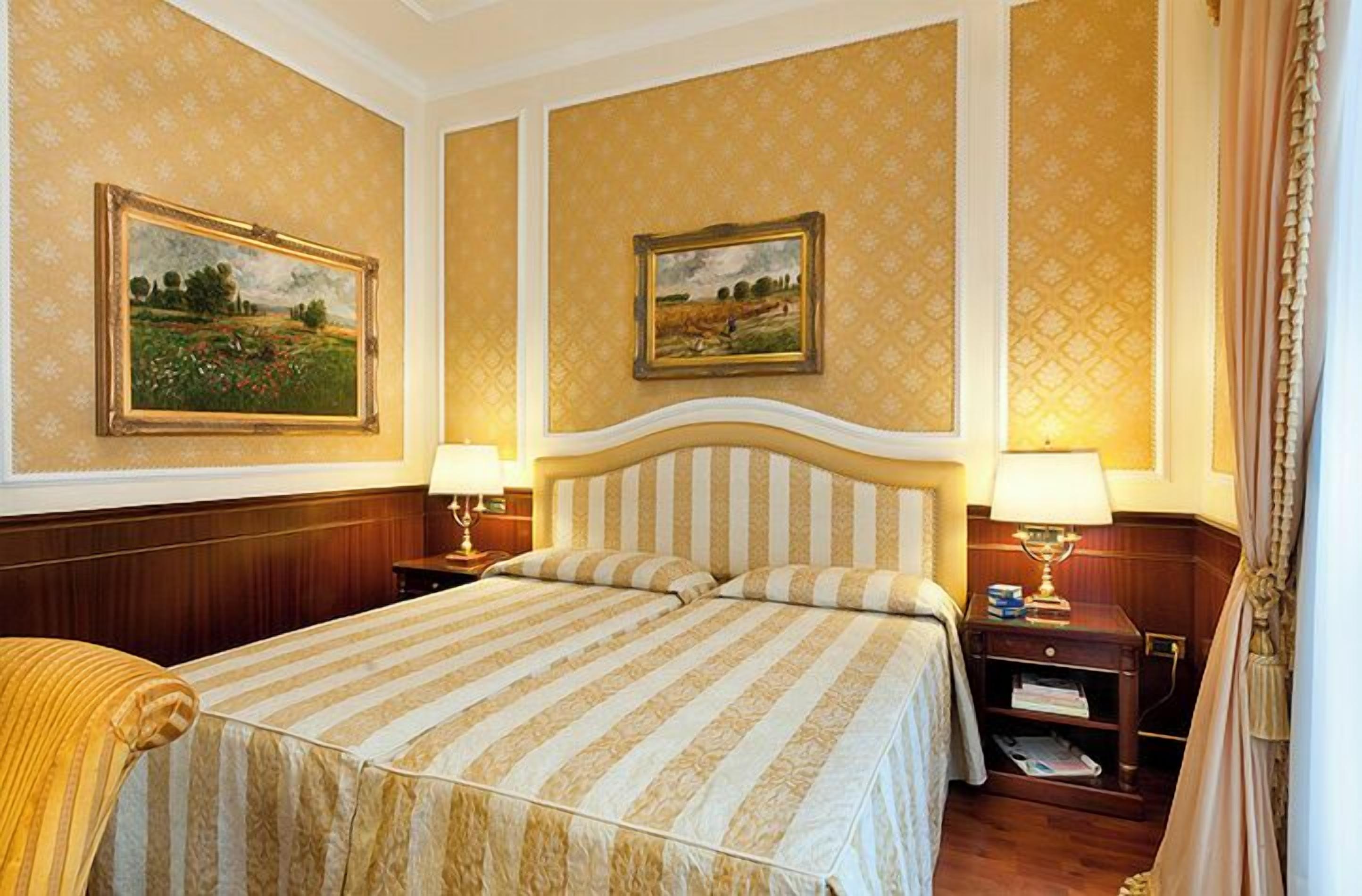 Habitació d'hotel amb un llit gran, dues taules de nit amb làmpades i dos quadres de paisatges emmarcats a les parets.