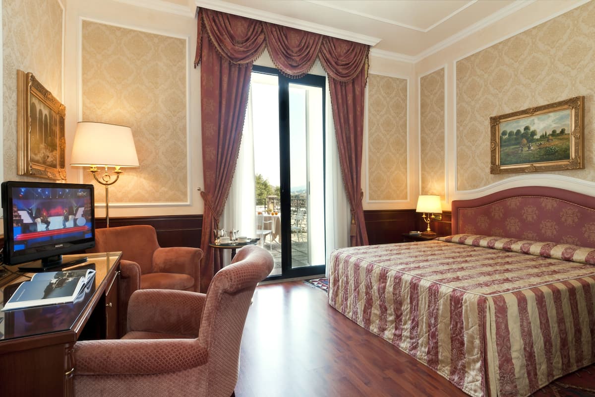 Quarto de hotel com cama, escrivaninha com computador, cadeira, televisão e um quadro na parede.