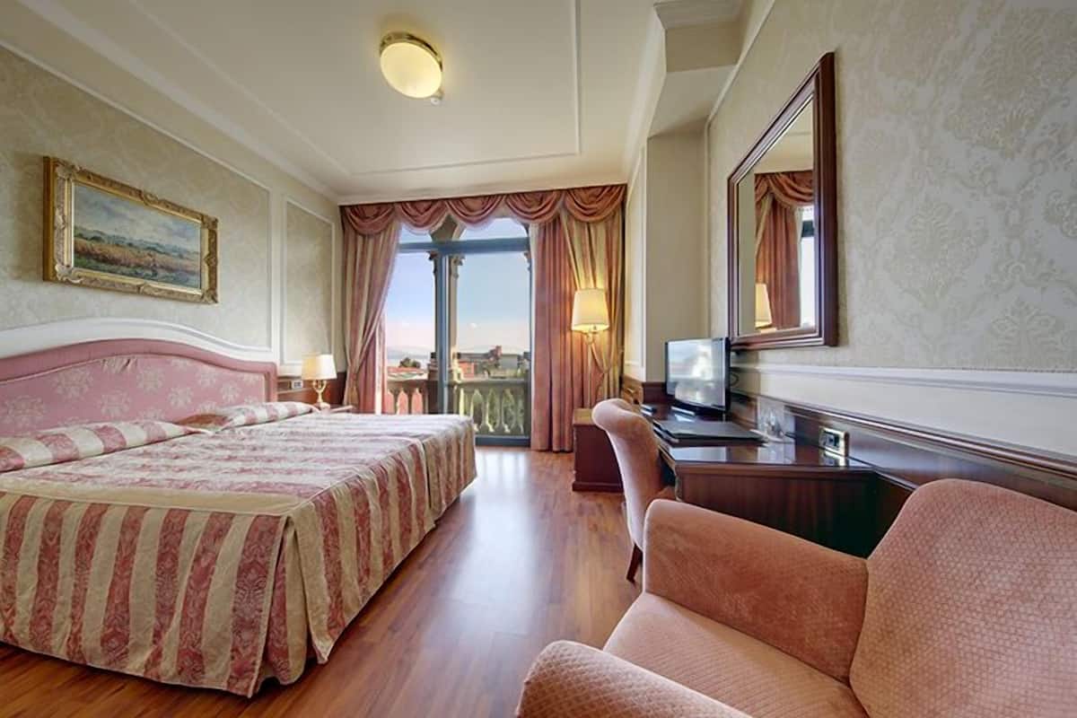 Quarto de hotel com uma cama grande, uma escrivaninha com televisão, uma cadeira e vista para o exterior por uma janela ampla.