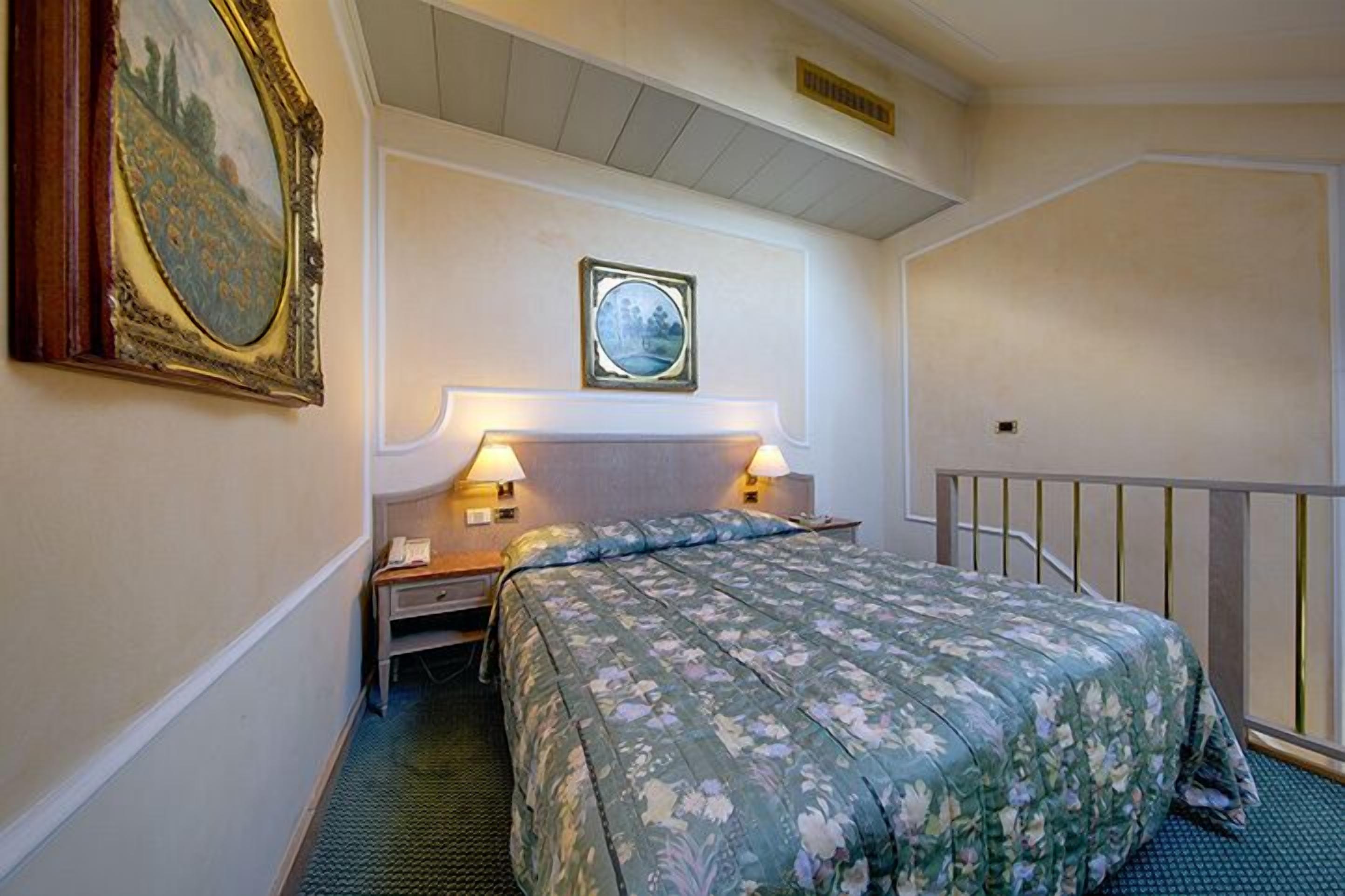 Habitación de hotel con cama, lámparas de noche, mesita de noche, un cuadro en la pared y una ventana pequeña.
