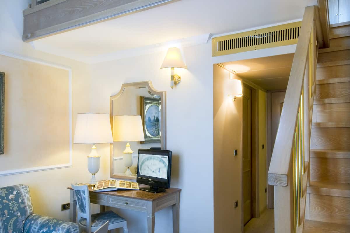 Uma sala de estar com escada de madeira, sofá, mesa de centro com livros e um laptop, e um corredor que dá para outro quarto.