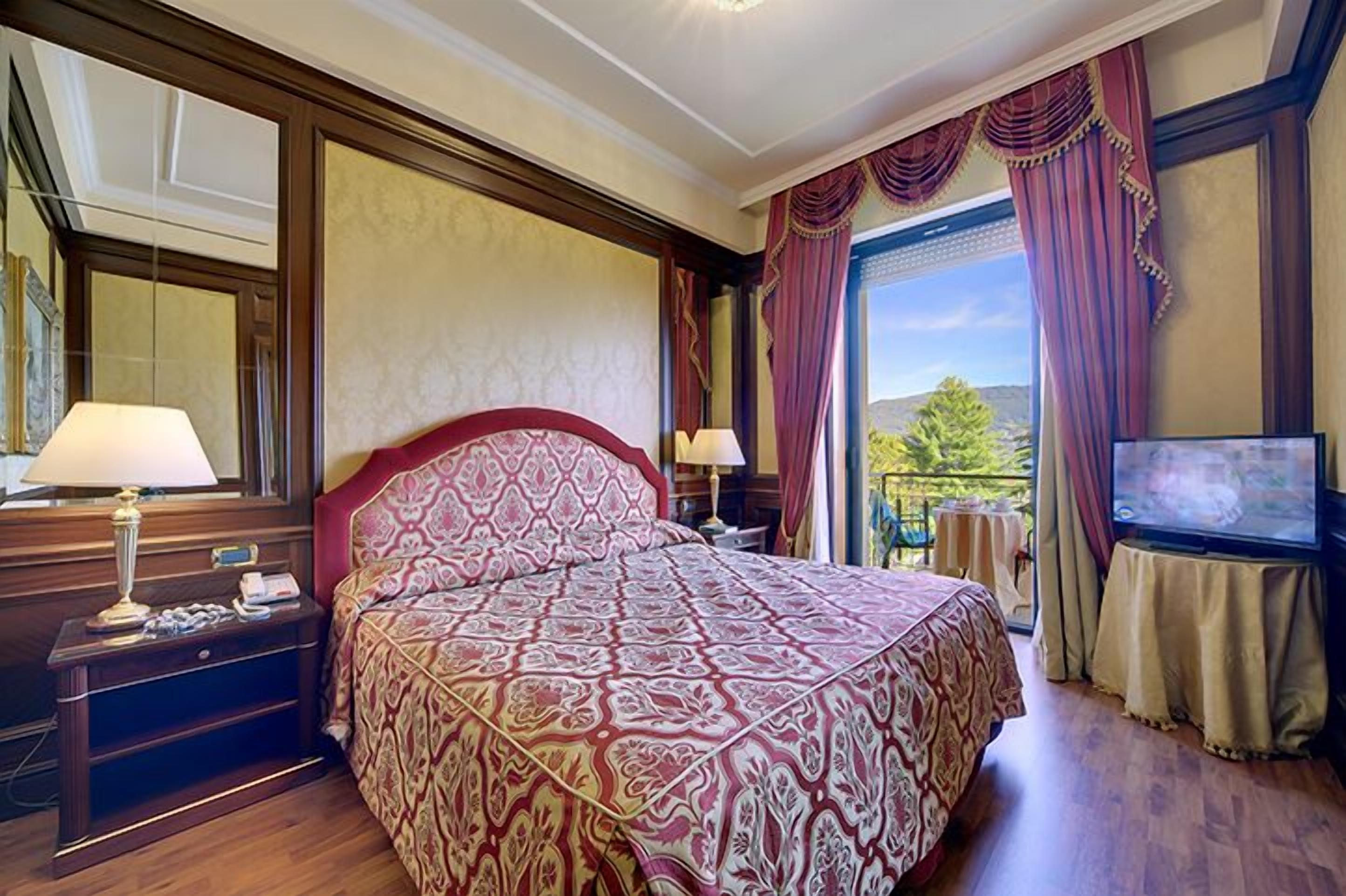 A hotel room with a large bed, two bedside tables with lamps, a television, and a balcony with a view of greenery.