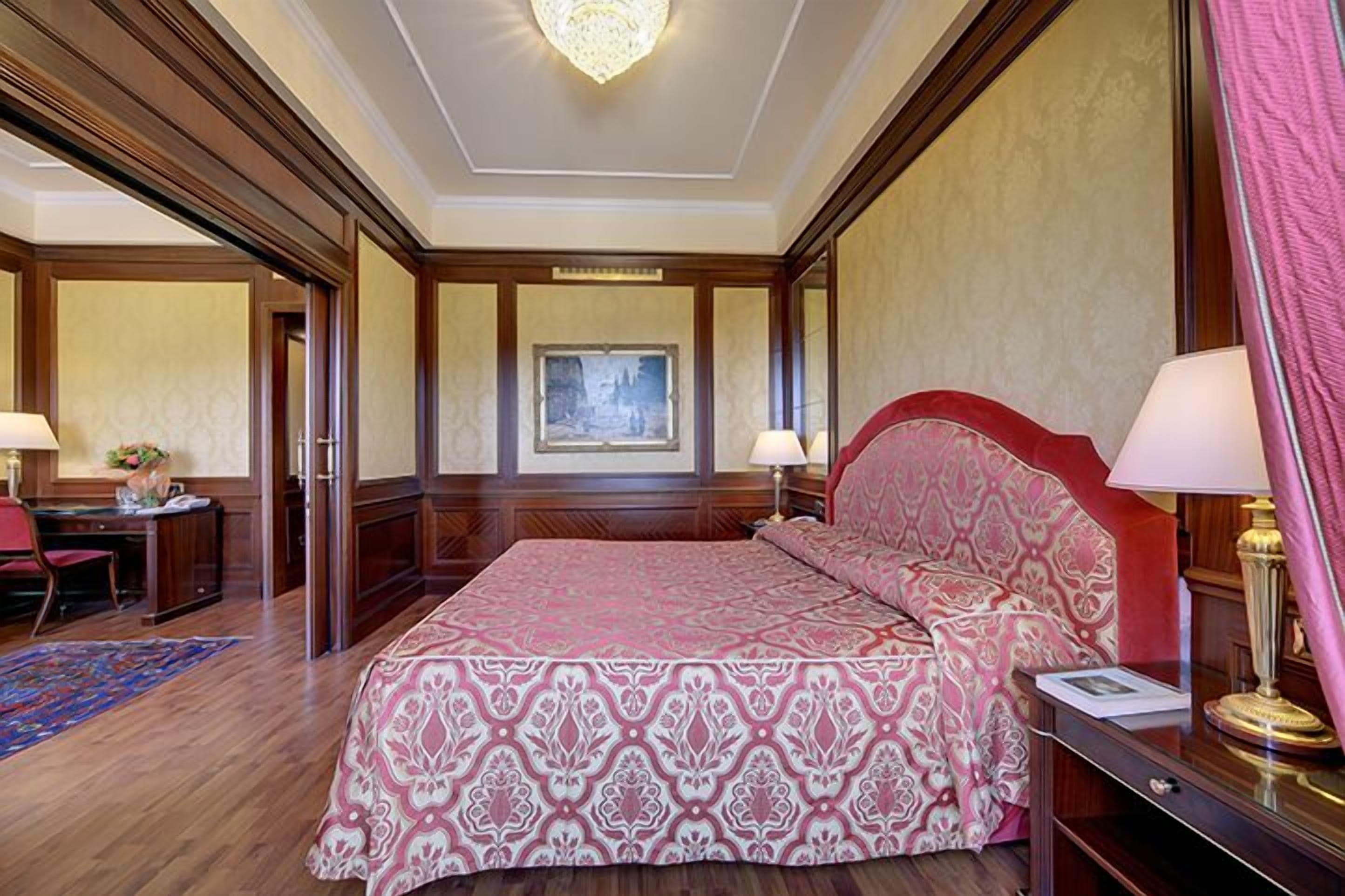 Une chambre d’hôtel avec un grand lit, des boiseries, un lustre et un bureau avec une chaise.