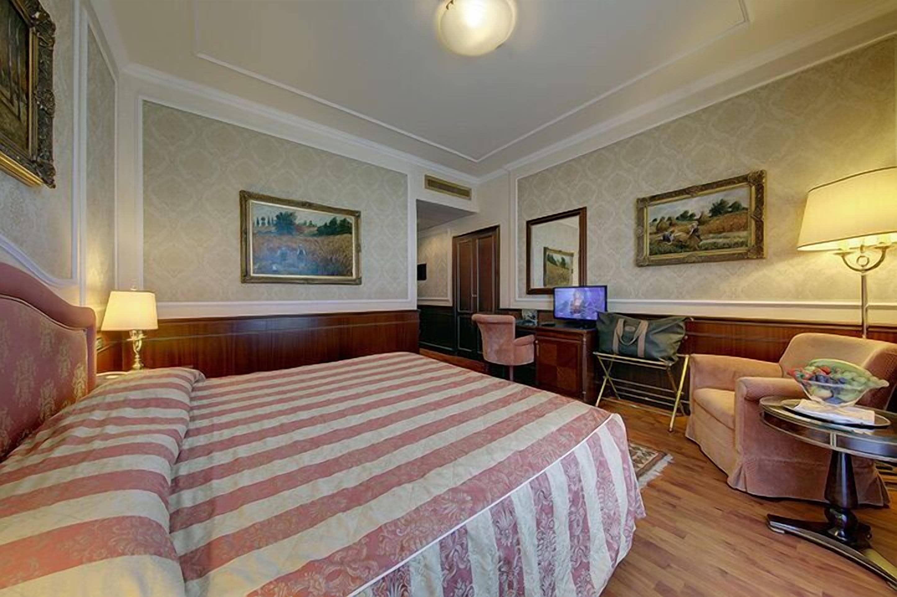 Una camera d'albergo con un letto grande, una scrivania con un computer, una sedia e due quadri appesi alle pareti.