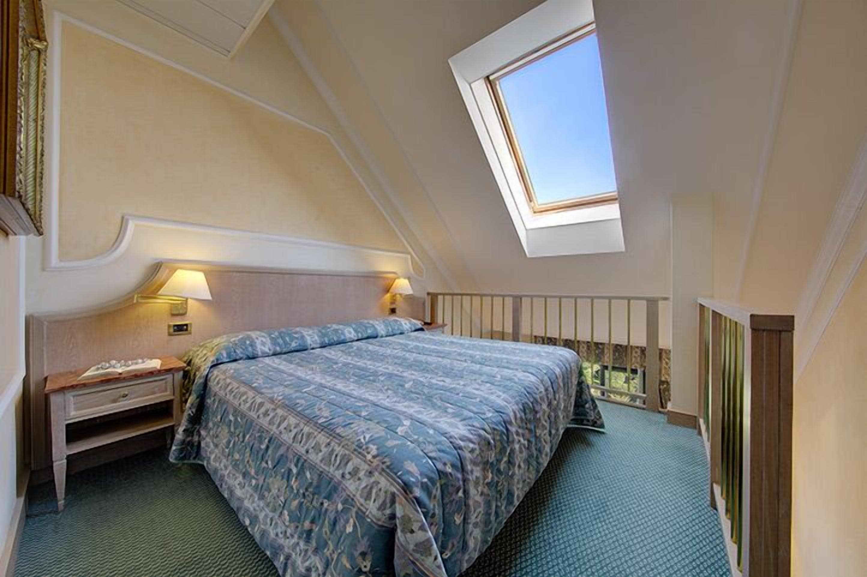 A bedroom with a skylight, a bed with a patterned bedspread, a nightstand, and a view of the outdoors.