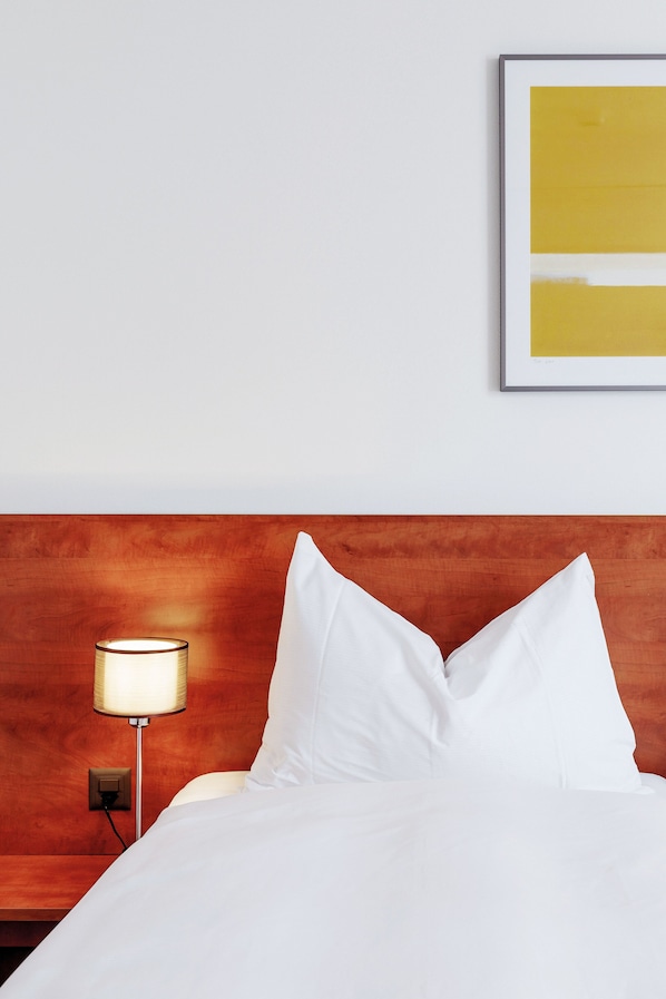 Habitación de hotel con cabecera de madera, cama blanca con almohadas, lámpara de noche y una obra de arte enmarcada en la pared.