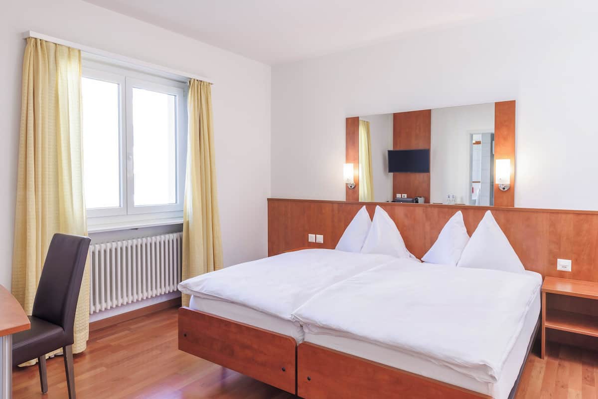 Habitación de hotel con cama de madera, un escritorio, una silla, una ventana con cortinas, un radiador y un televisor fijado a la pared.