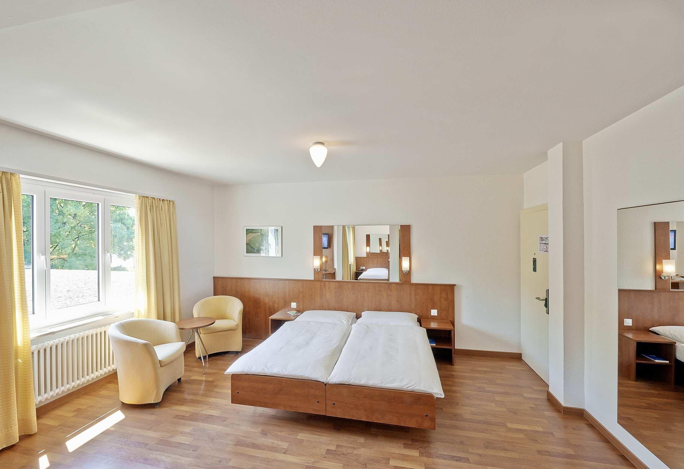 Ett hotellrum med en dubbelsäng, ett litet bord, två stolar, ett fönster med gardiner, ett element och en spegel på väggen.