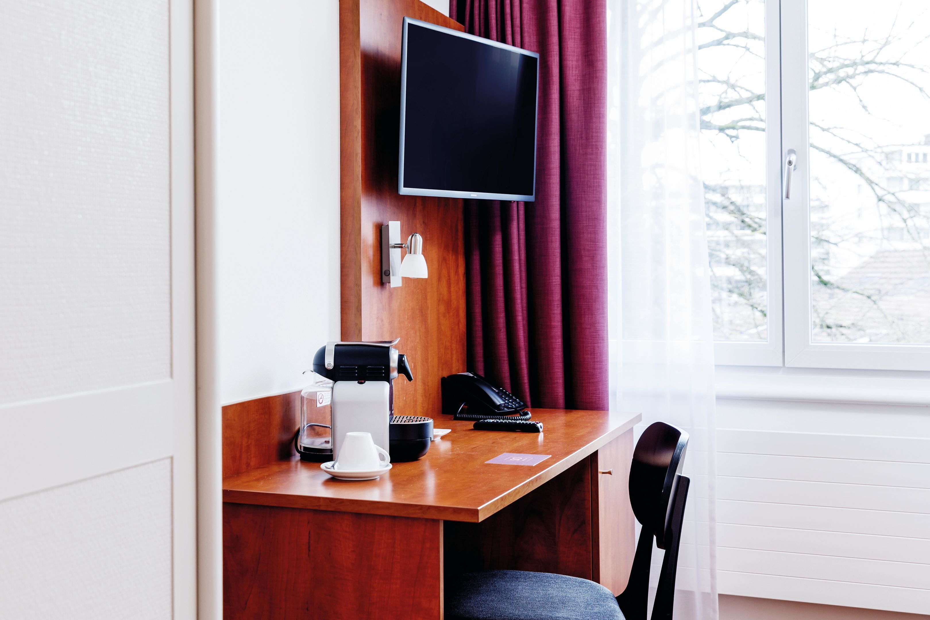 Habitación de hotel con un escritorio de madera, un televisor montado en la pared, cafetera, teléfono y una silla.