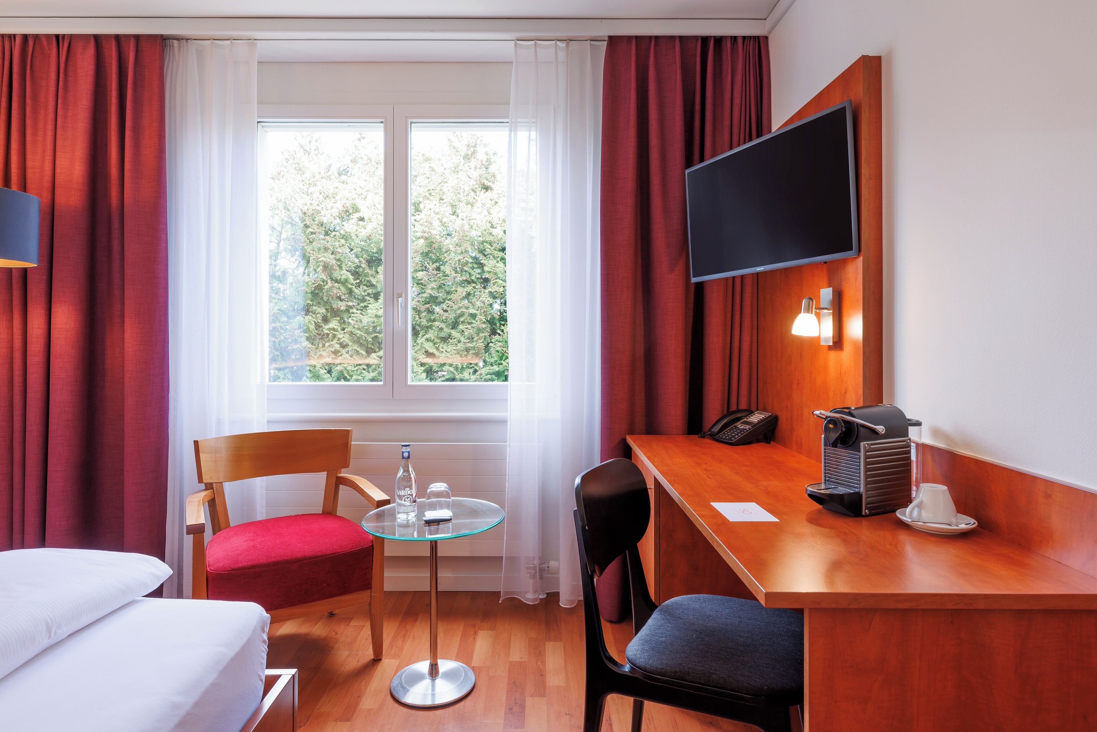 A hotel room with a bed, a desk with a coffee maker, a TV, and a window with curtains.
