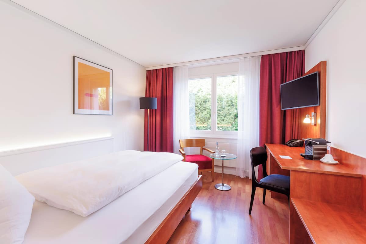 Habitación de hotel con cama, escritorio, silla, televisor y ventana con cortinas.