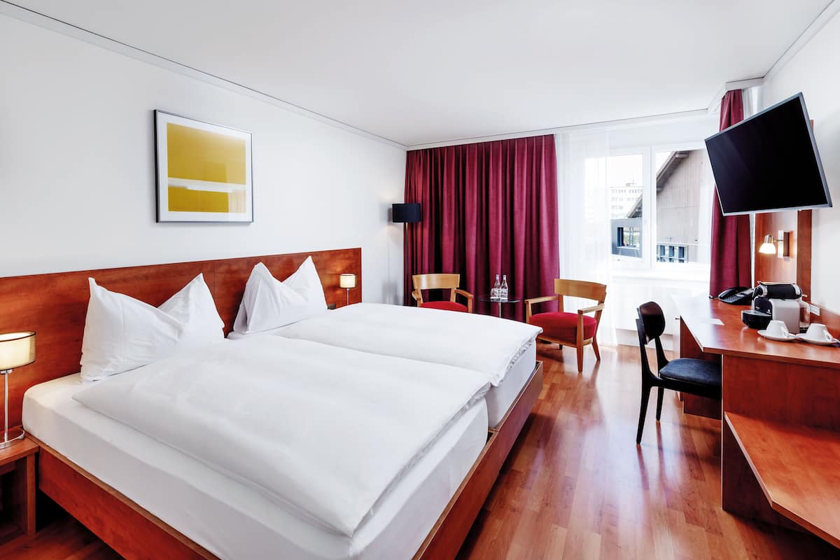 Habitación de hotel con una cama grande, un escritorio, una silla, un televisor y una ventana con cortinas.