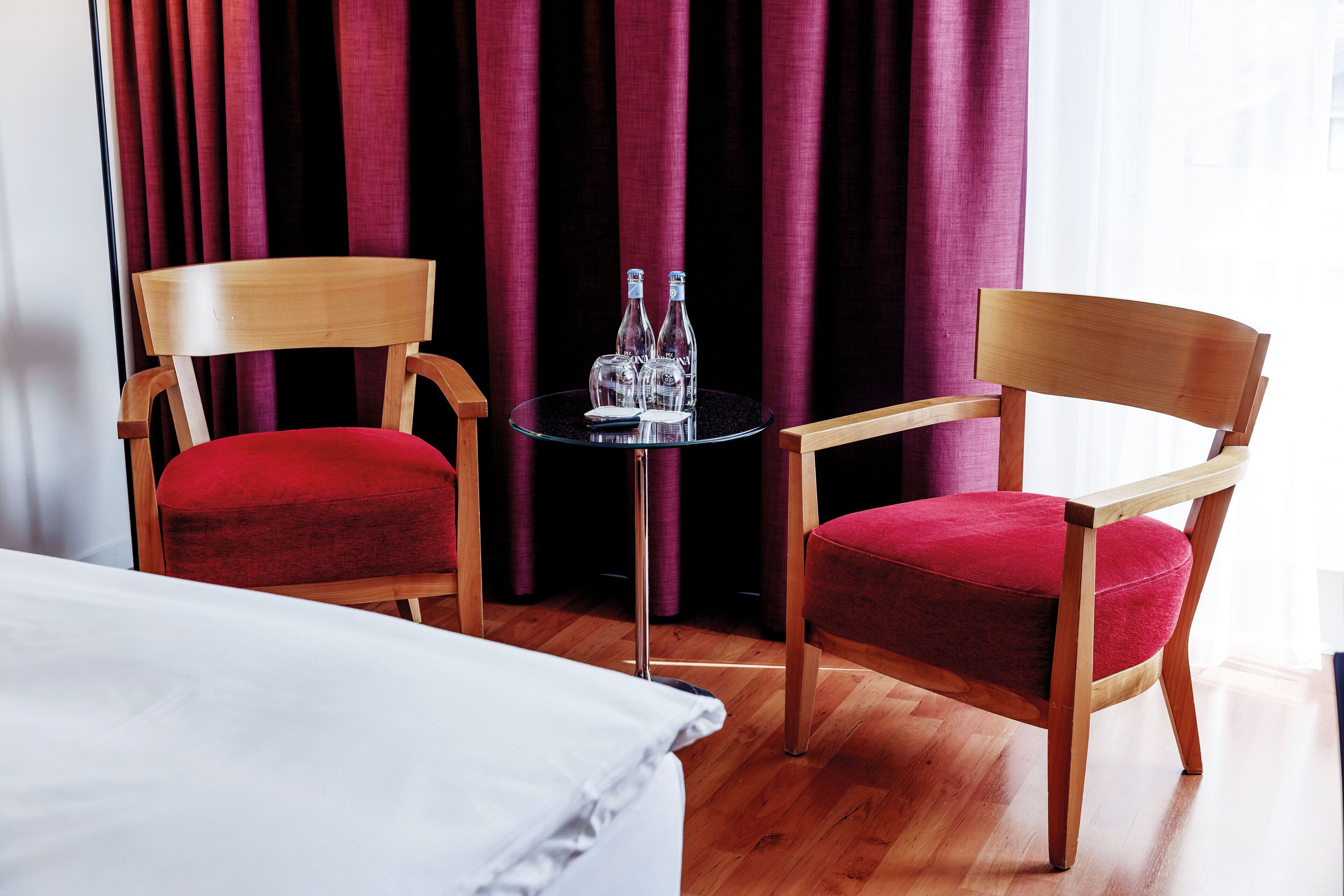 Een hotelkamer met twee houten stoelen met rode kussens, een klein rond tafeltje met twee flessen en een bed met witte lakens.