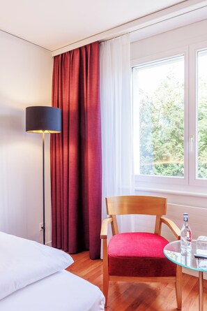 Une chambre d’hôtel avec un lit, une chaise, une table avec une bouteille et des verres, une lampe sur pied et une fenêtre avec des rideaux.