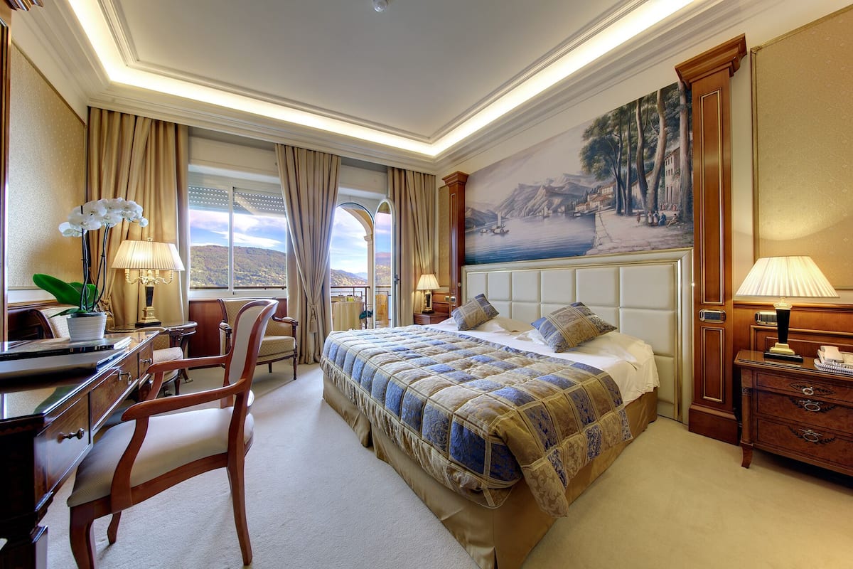 A hotel room with a large bed, a desk, a chair, a lamp, and a painting on the wall.