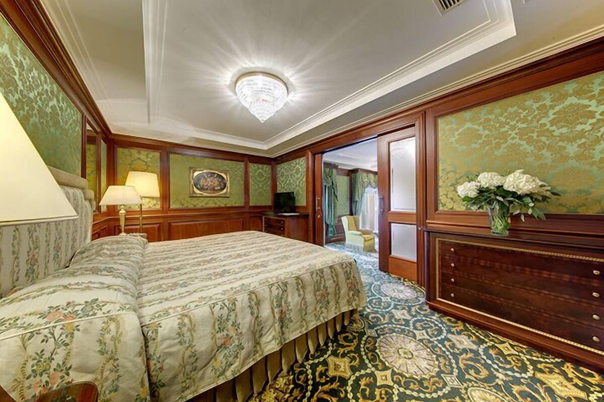 A traditional hotel room with a large bed, a wooden dresser, a chandelier, and patterned wallpaper.