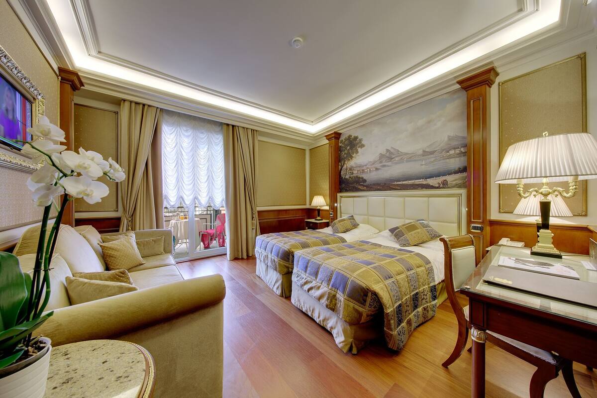 A hotel room with two beds, a sofa, a desk, a lamp, and a painting on the wall.