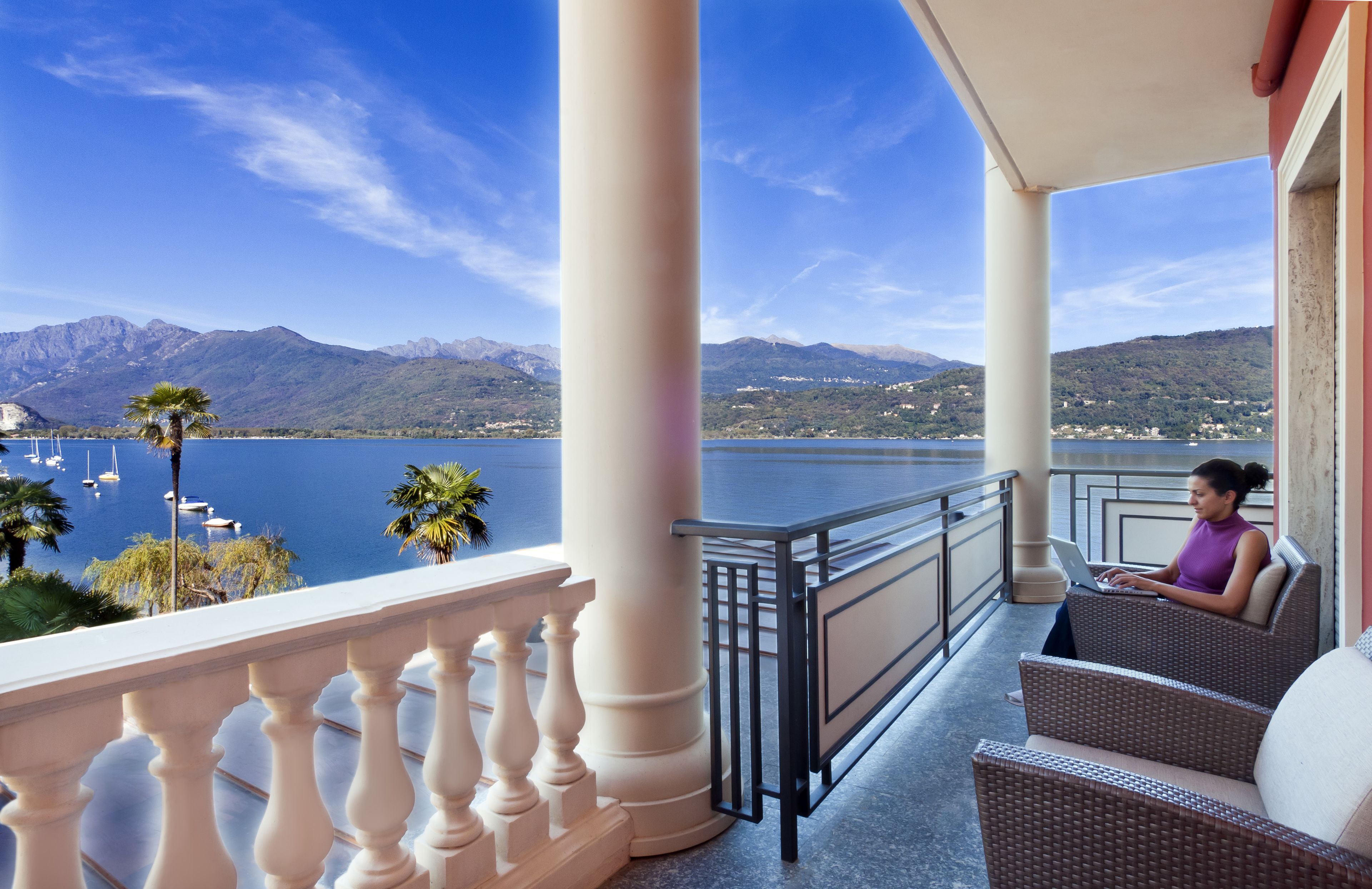 Un balcon avec vue sur un lac et des montagnes, un canapé en osier et une personne utilisant un ordinateur portable.