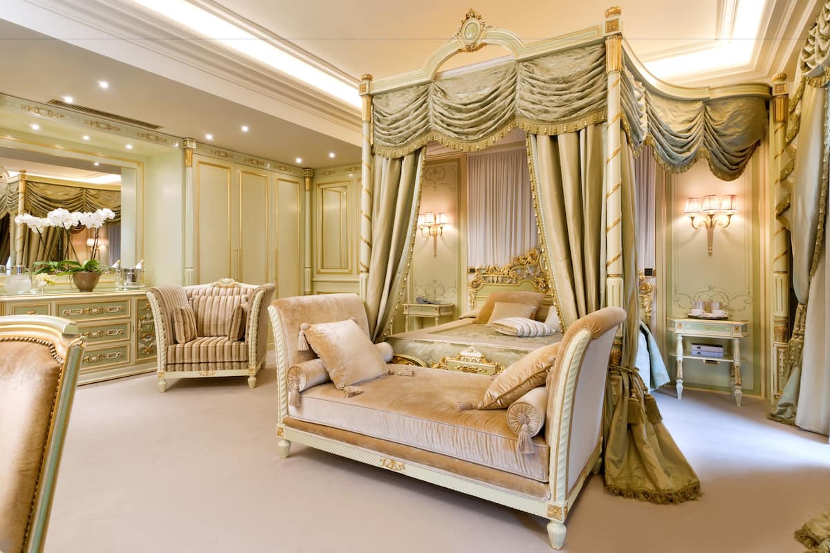 A luxurious bedroom with a four-poster bed, a chaise lounge, and a vanity with a mirror.