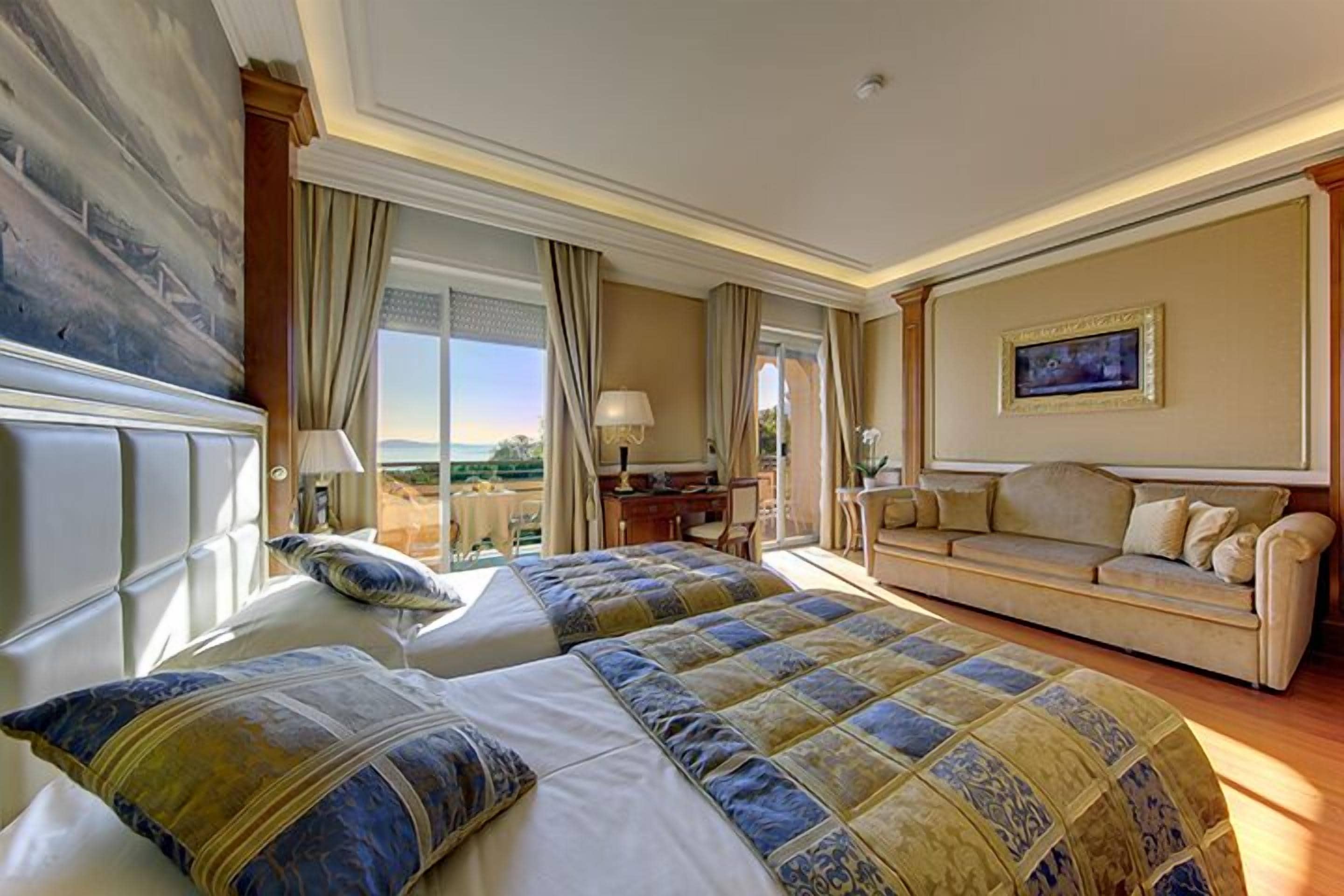 A hotel room with two beds, a sofa, a desk, and a large window with a view.