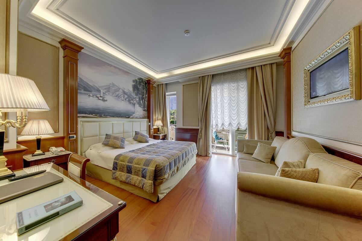 A hotel room with a large bed, a sofa, a desk with a lamp, and a painting on the wall.