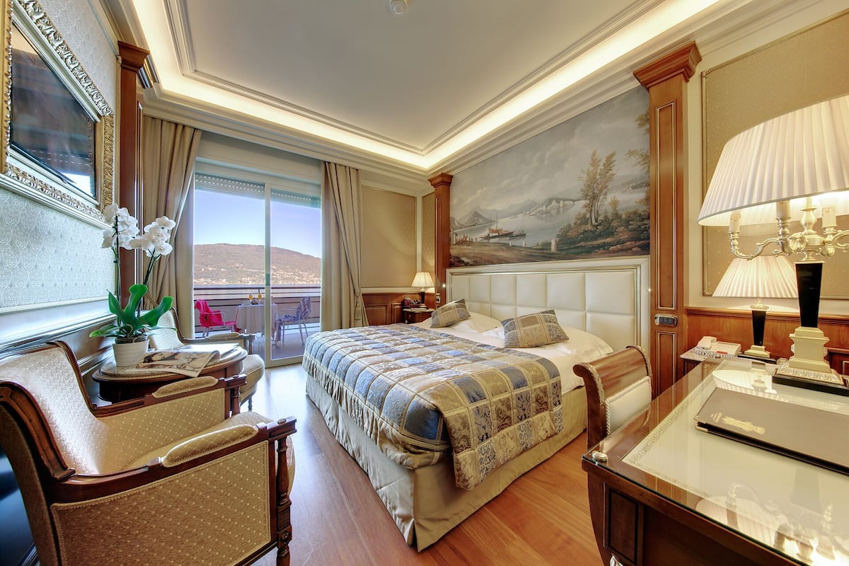 A spacious hotel room with a large bed, a sitting area with a chair and table, a balcony with a view of the landscape, and a painting on the wall.