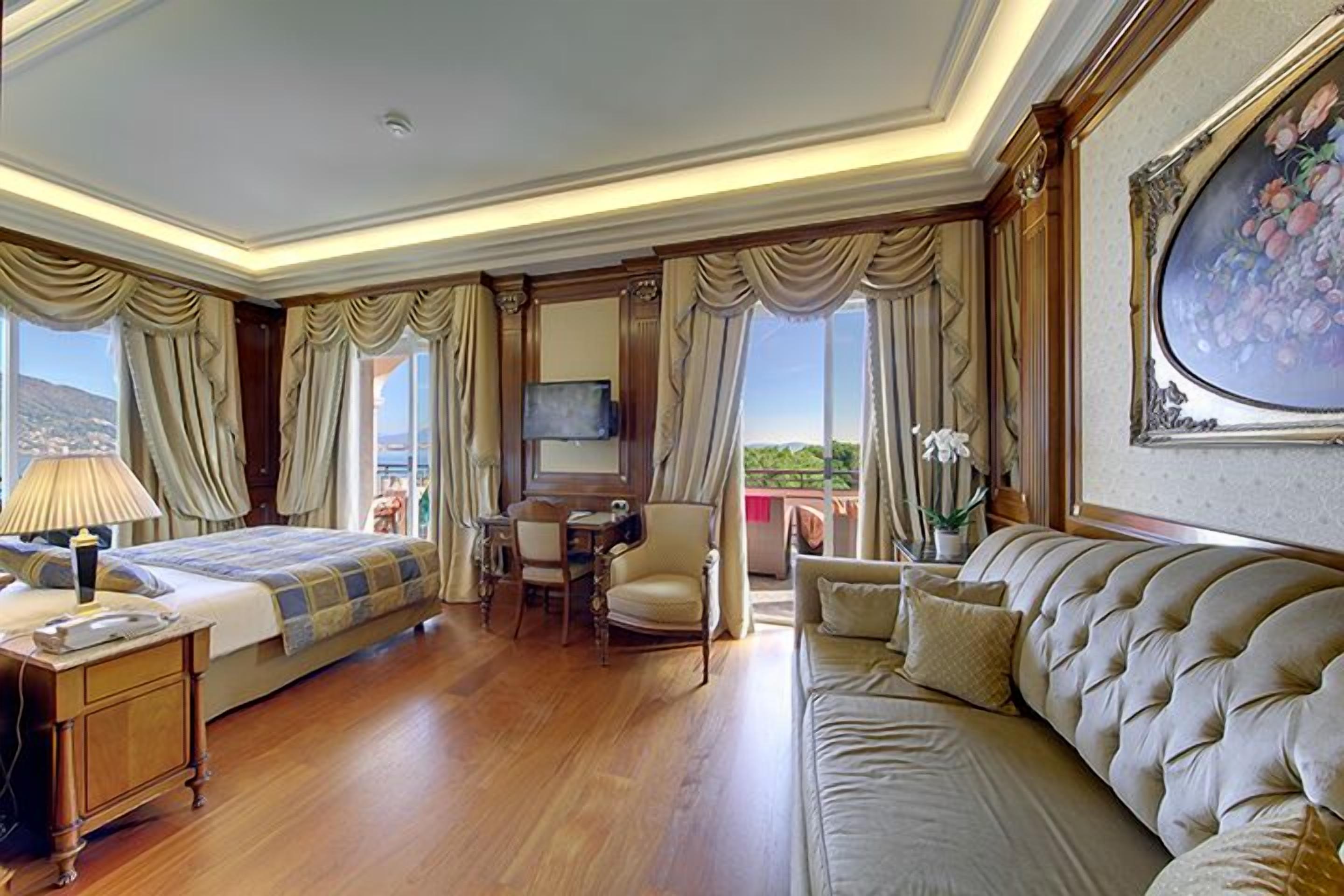 A hotel room with a bed, a sofa, a desk with a chair, a TV, and a large window with curtains.