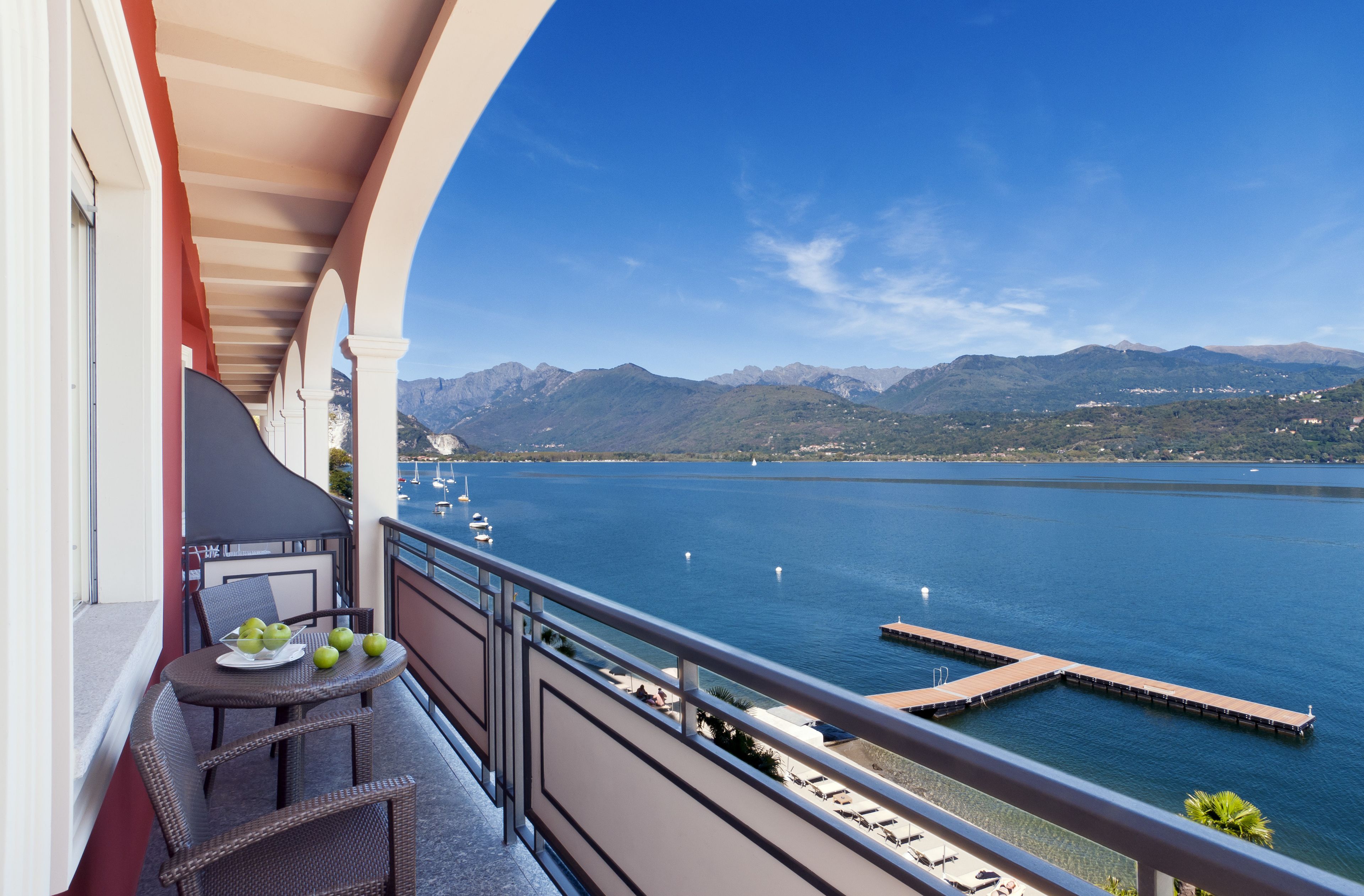 Un balcon agrémenté de meubles en osier, une table avec des pommes, et une vue sur un lac avec des montagnes en arrière-plan.