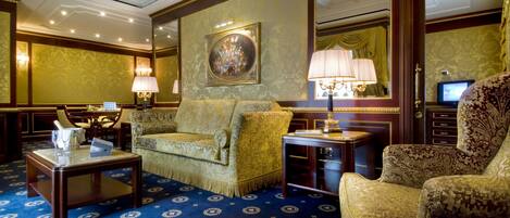 A luxurious room with a patterned carpet, a sofa, a chair, a small table with a lamp, and a television.