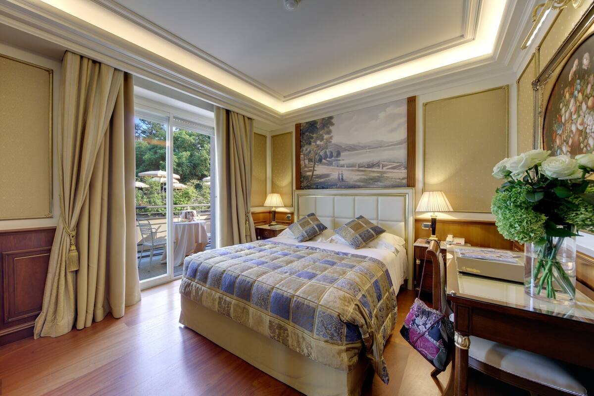 A hotel room with a large bed, two bedside tables with lamps, a painting on the wall, and a view of the outdoors through the sliding glass door.