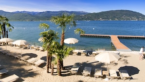 A beach with sun loungers and umbrellas, a wooden pier extending into the water, and mountains in the background. - Hotel Splendid (Baveno)