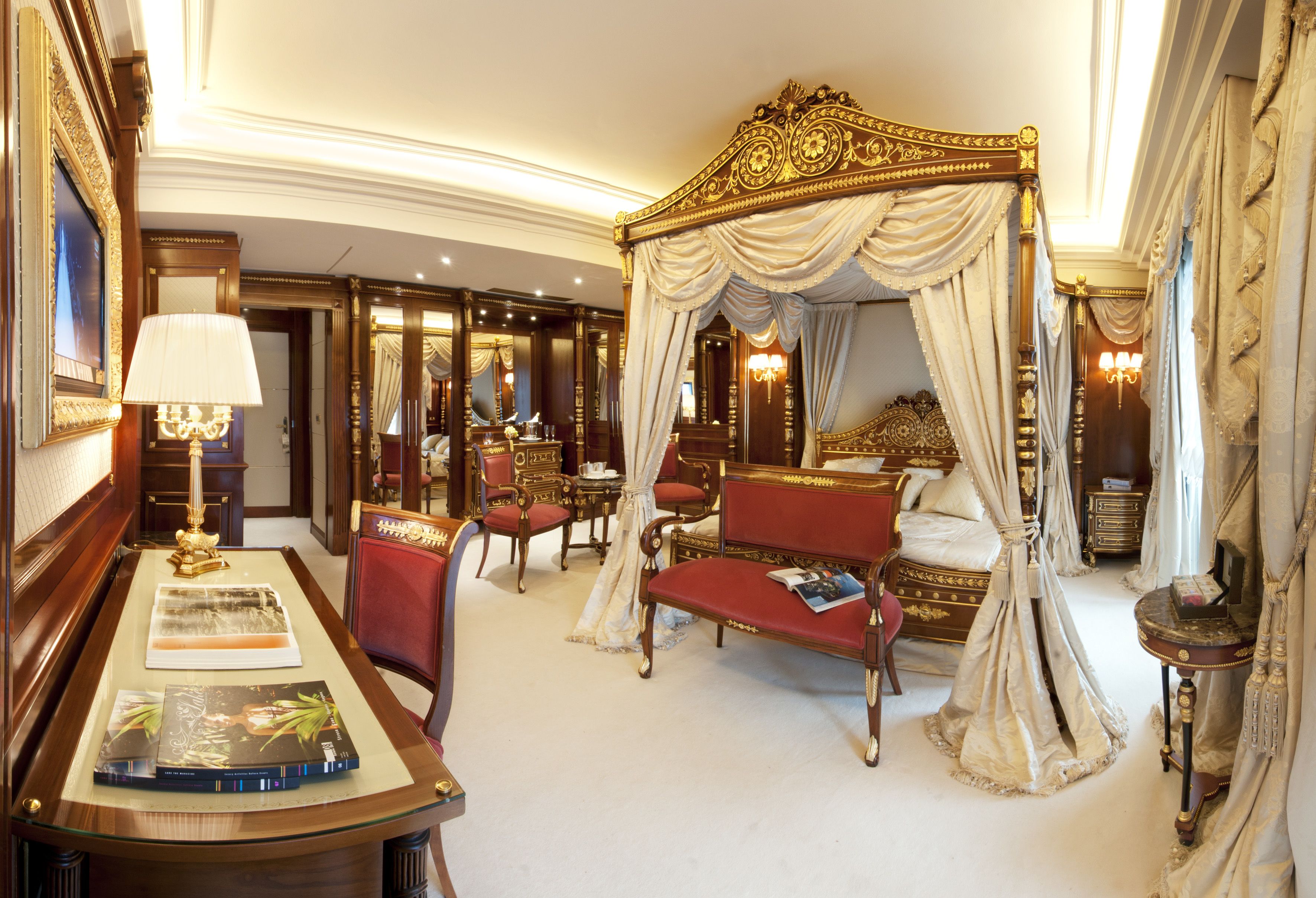 Een luxueuze slaapkamer met een hemelbed, een bureau met lamp en een rode fauteuil.