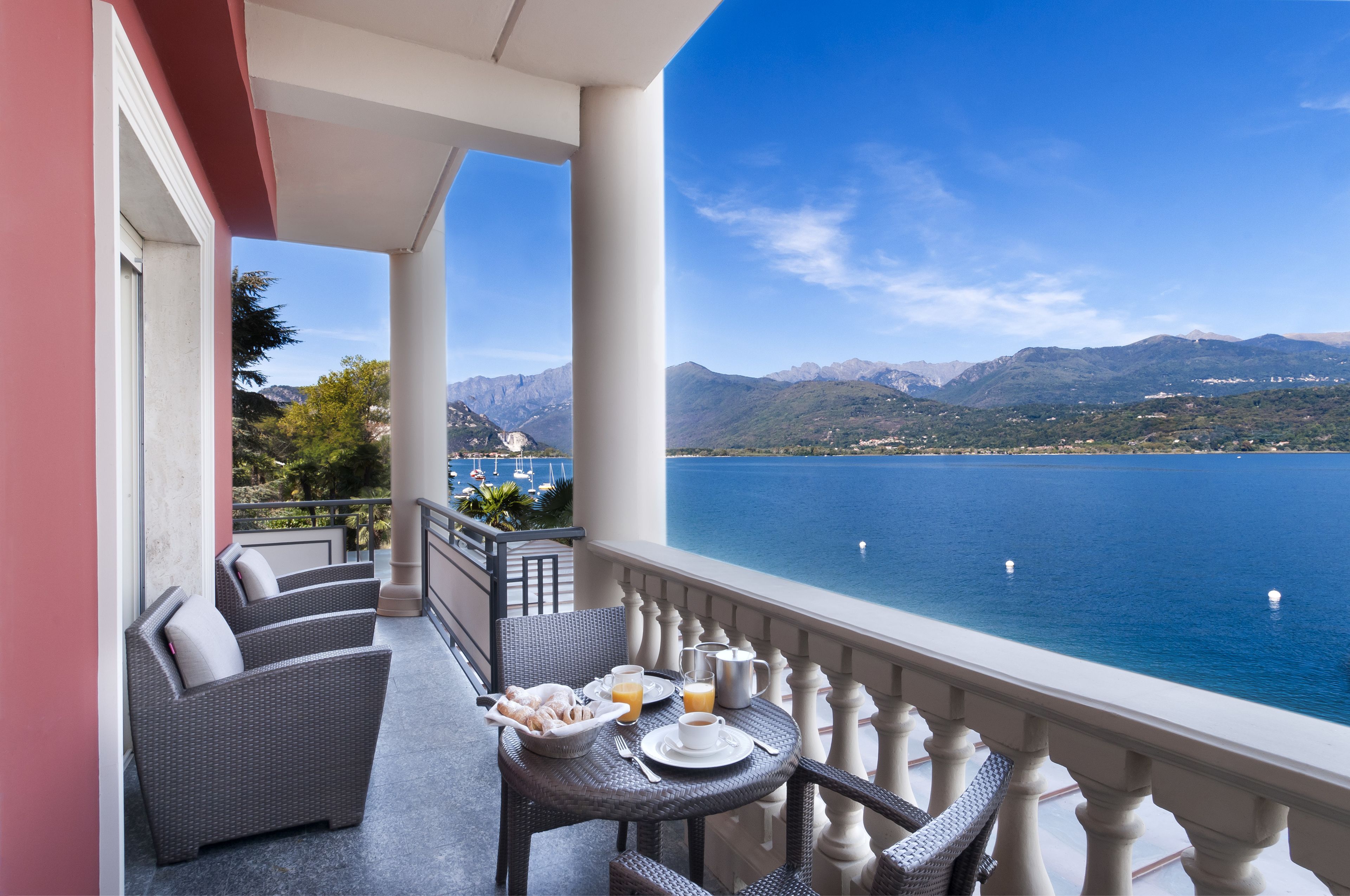 Ein Balkon mit Korbmöbeln, einem gedeckten Frühstückstisch und Blick auf einen See und Berge.