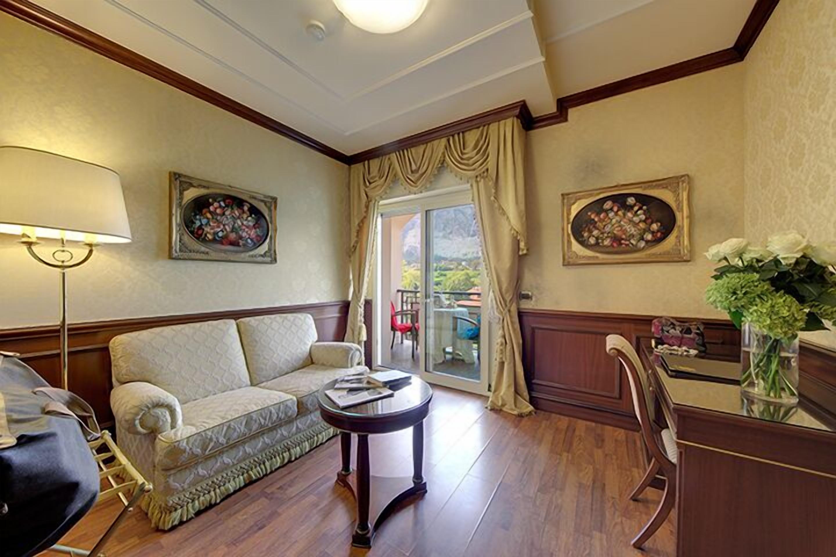 A hotel room with a sofa, a small round table, a desk with a chair, and a view through a glass door to the outside.