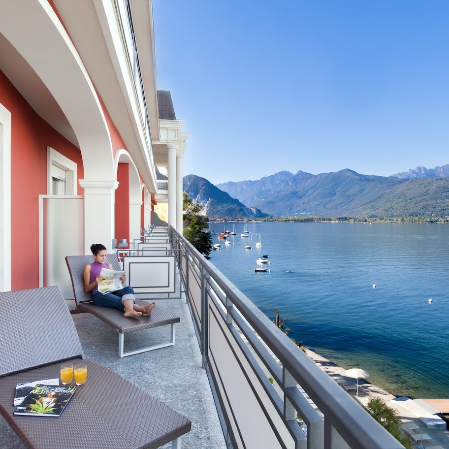 A balcony with a table and chairs, overlooking a lake with boats and mountains in the background.