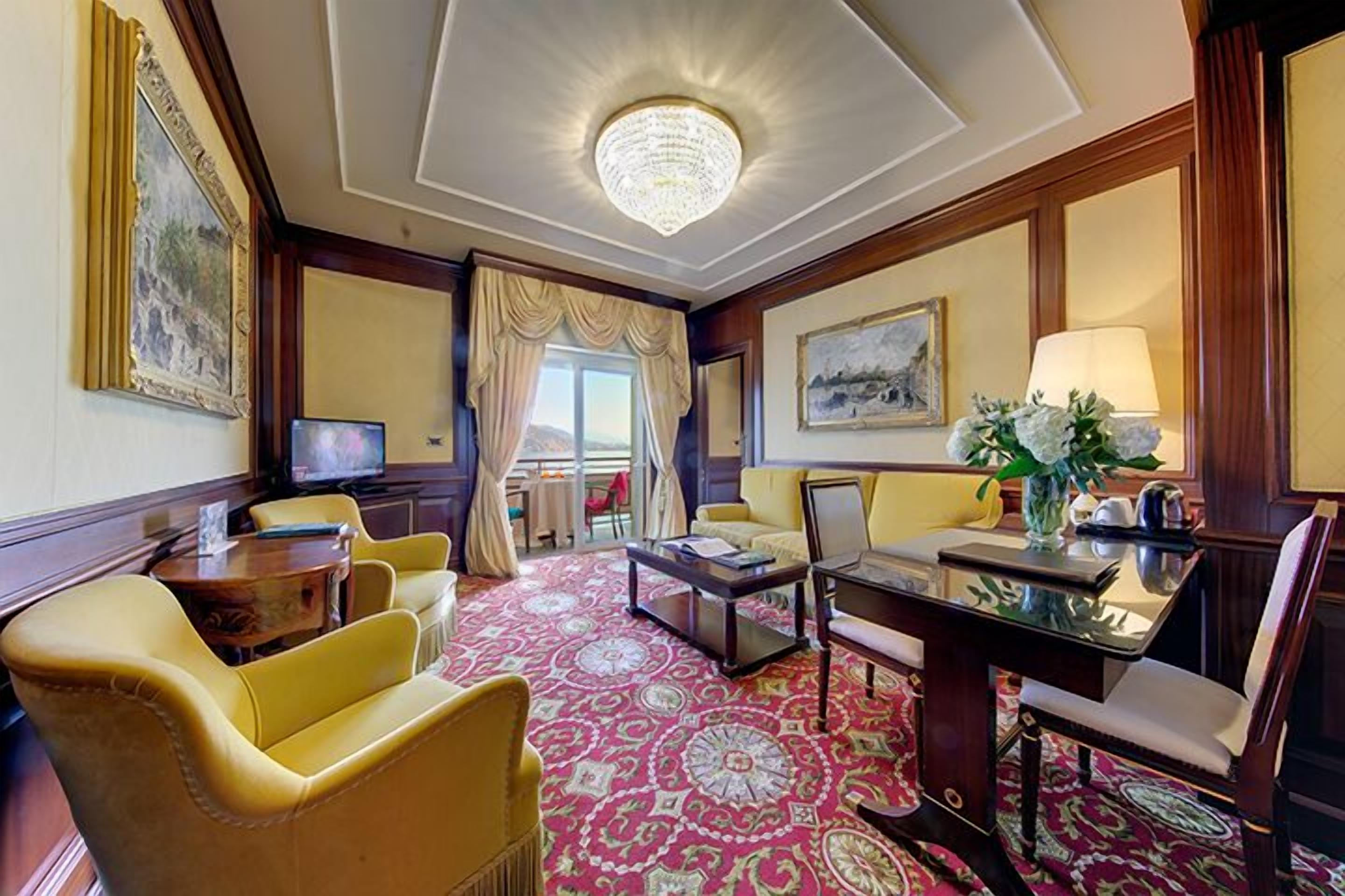Una camera d'albergo con un lampadario grande e decorativo, una moquette a motivi e un'area salotto con poltrone gialle e un tavolo.
