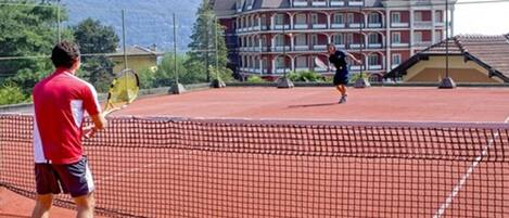 A tennis court with a net in the middle, surrounded by a fence. In the background, there is a large building with multiple balconies.