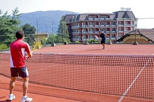 Una cancha de tenis con una red en el centro, rodeada por una cerca. En el fondo, se observa un edificio grande con varios balcones.