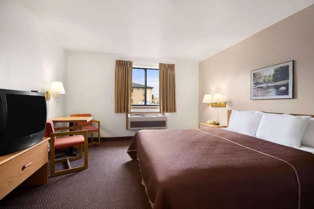 A hotel room with a bed, a TV, a desk with a chair, a window with curtains, and a lamp.