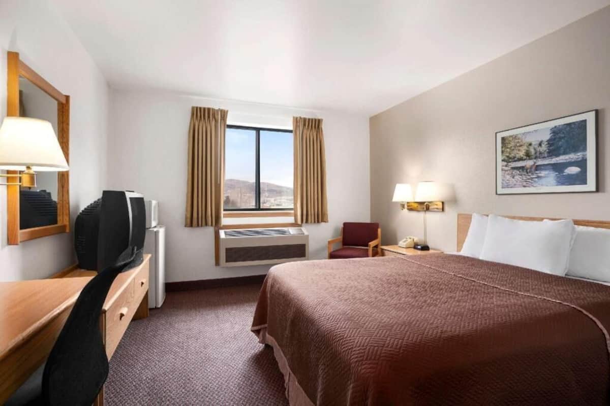 A hotel room with a bed, desk, chair, window with curtains, and a painting on the wall.