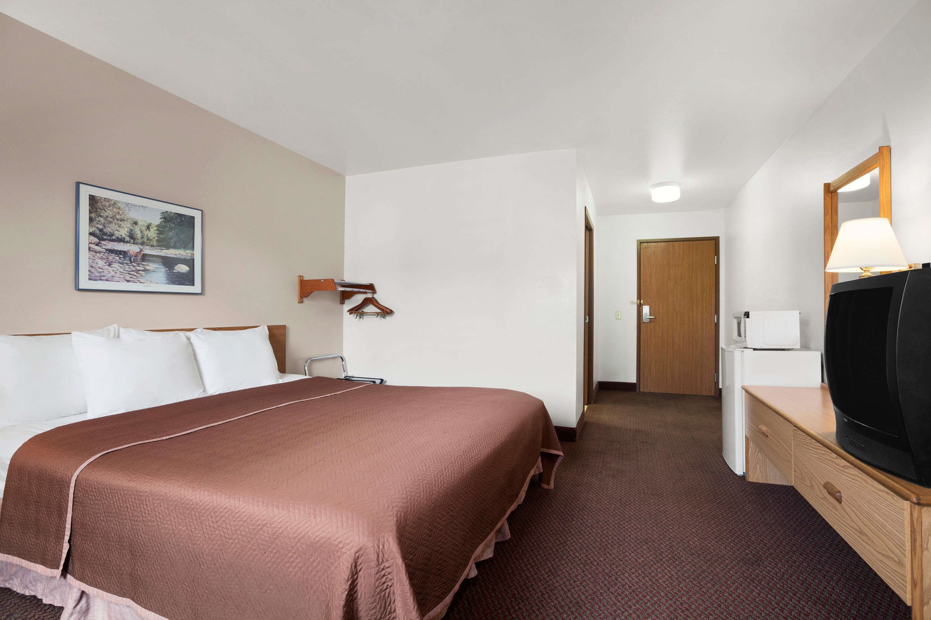 A hotel room with a large bed, a television, a desk with a lamp, and a door leading to another room.
