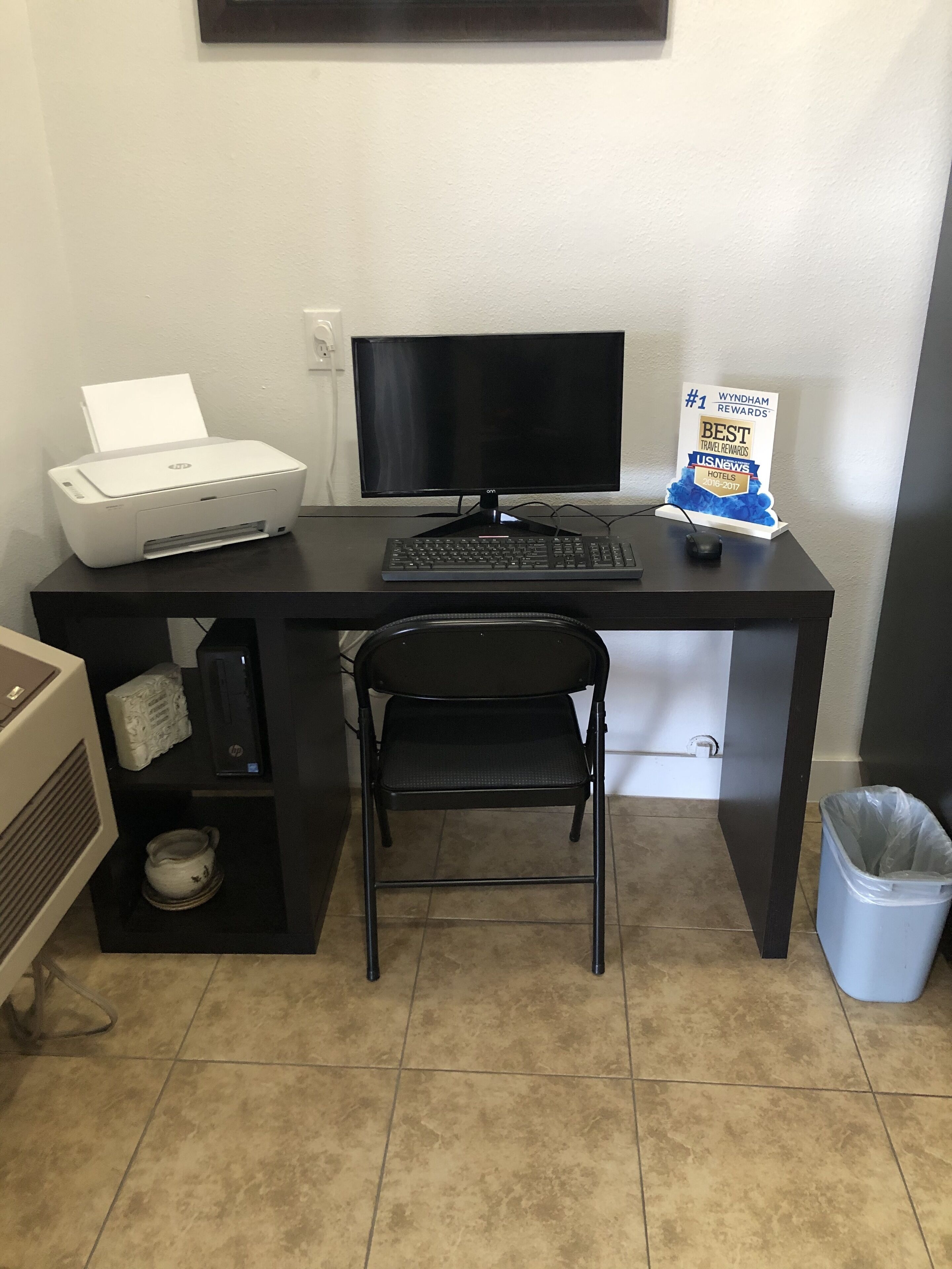 A compact office space with a desk, a printer, a monitor, a keyboard, a chair, a trash bin, and a sign indicating Wyndham Rewards.