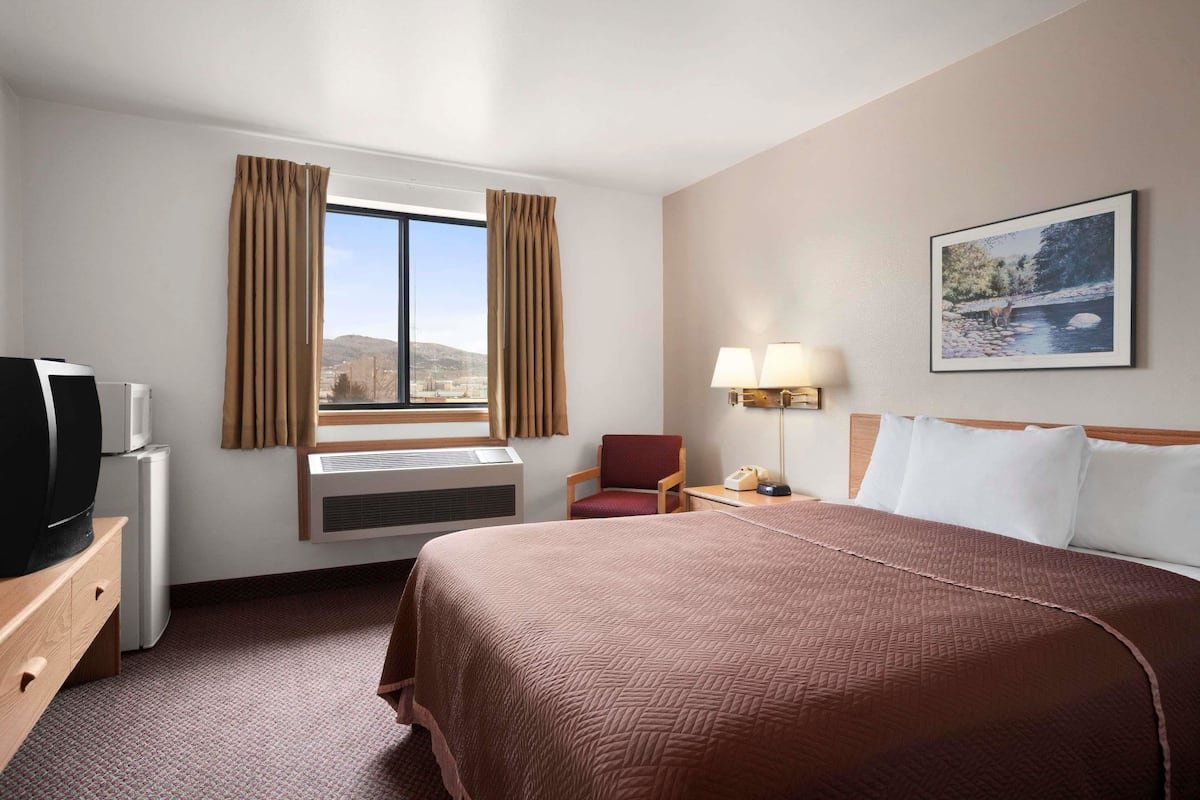 A hotel room with a large bed, a TV, a chair, a desk with a lamp, and a window with curtains.