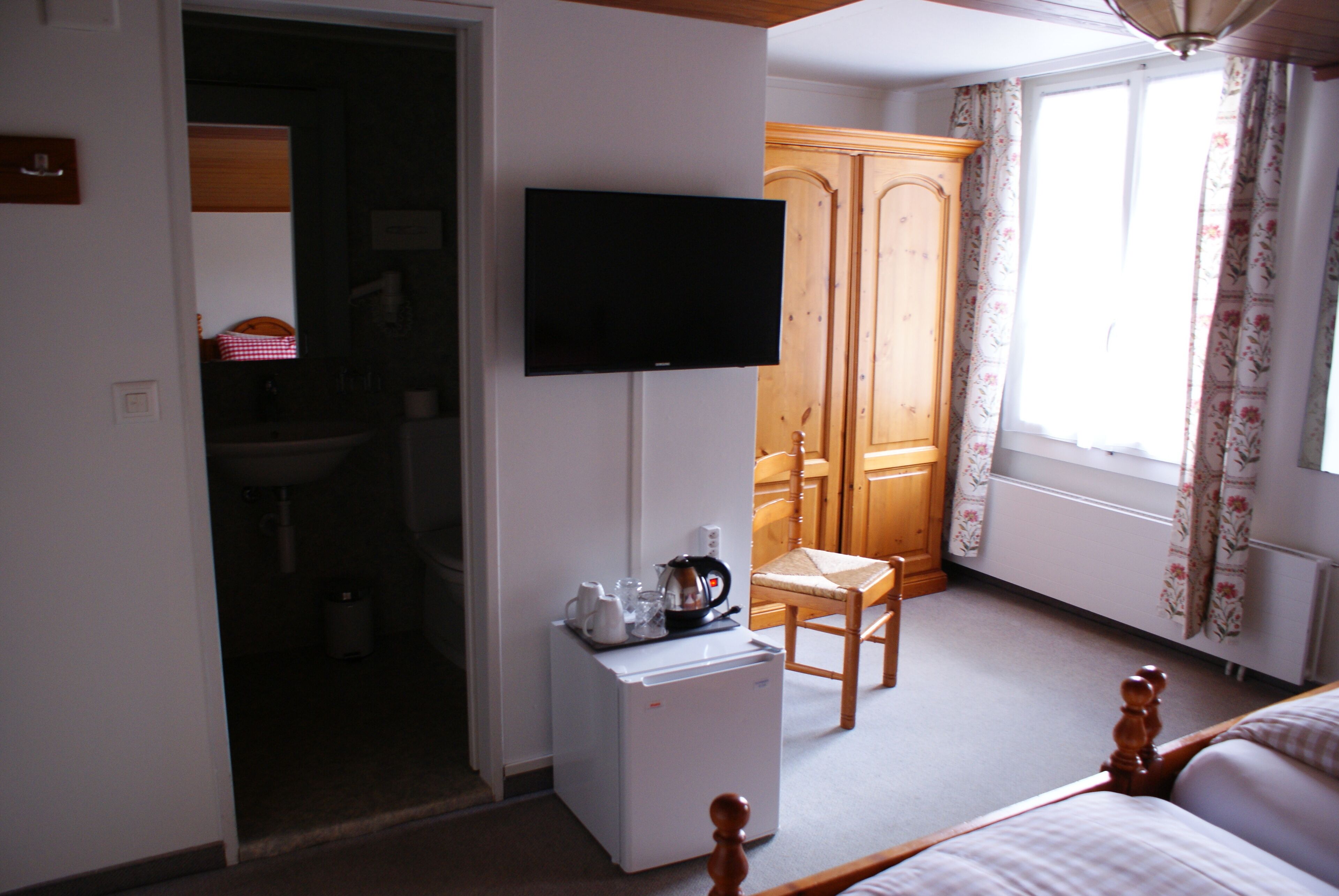 A hotel room with a bed, a television mounted on the wall, a small refrigerator, a wooden chair, and a wardrobe.