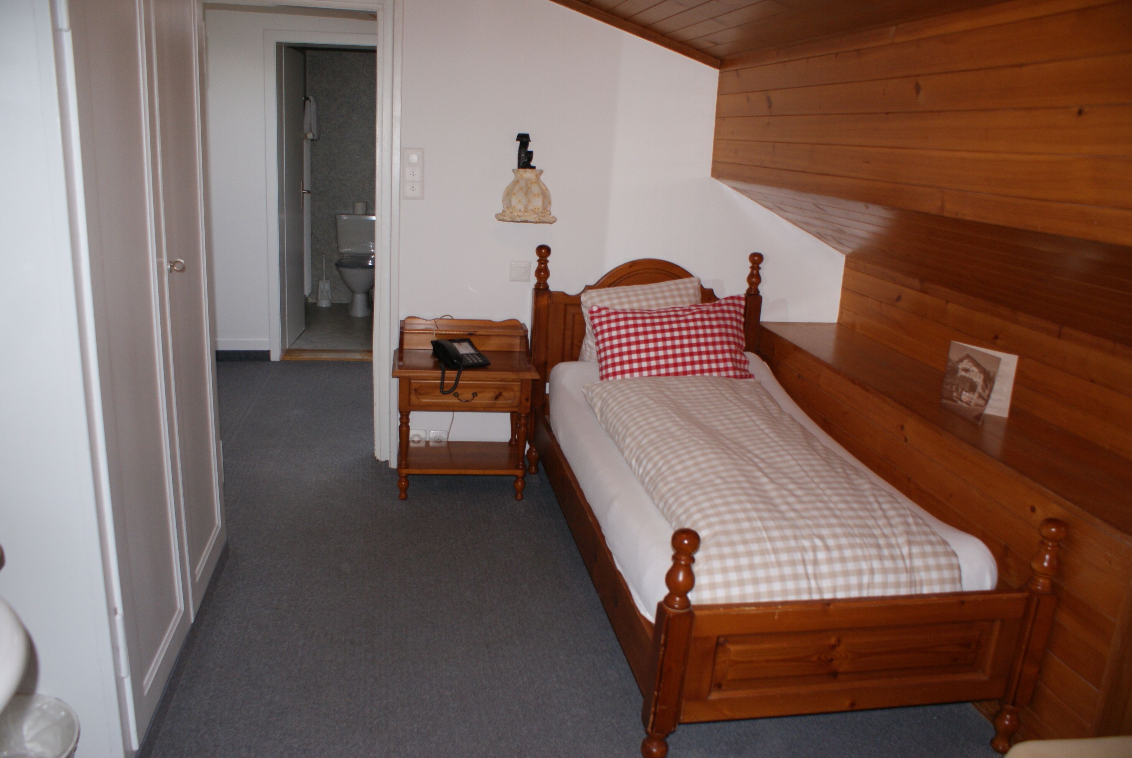 Un dormitorio pequeño y ordenado con una cama de madera, una mesita de noche con lámpara y vistas al baño.